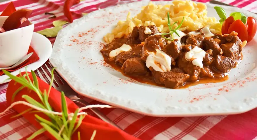 En smak av mat i Budapest: mustig gulasch med nokedli, garnerad med gräddfil och en rosmarinkvist. En röd chili och en tomatblomma fulländar upplägget.