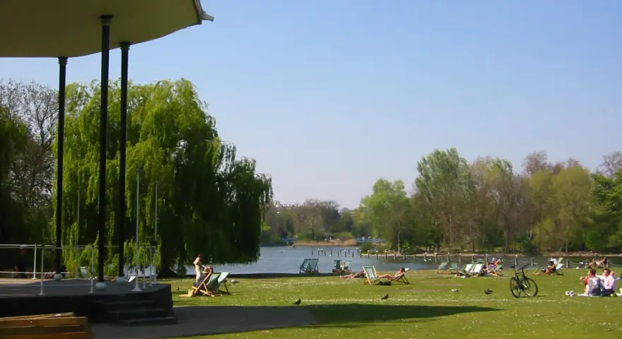En solig dag i en park i London. Människor vilar på solstolar vid en sjö, med en grönskande trädgård och en paviljong i förgrunden.