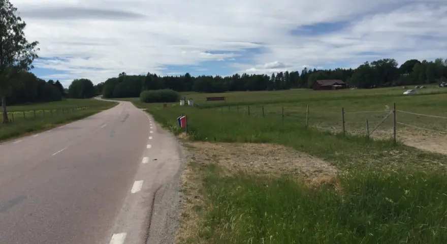 Vacker väg kantad av grönska, perfekt för att utforska Sveriges cykelleder. En röd lada skymtar i bakgrunden.