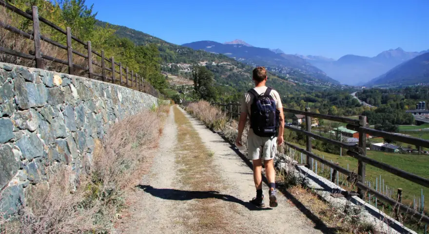Man vandrar i Italien på en grusväg med stenmur och staket. Vacker bergig dal i bakgrunden.