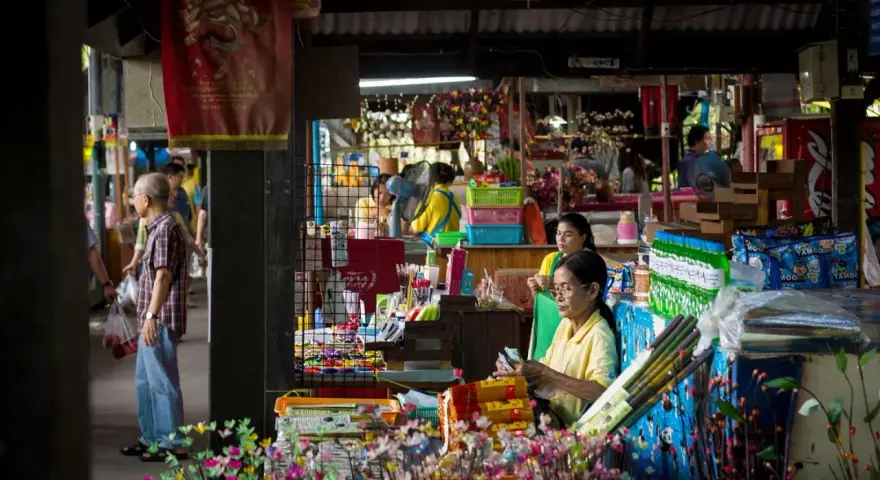 En livlig marknad i Bangkok med försäljare och kunder bland färgglada varor och blommor.