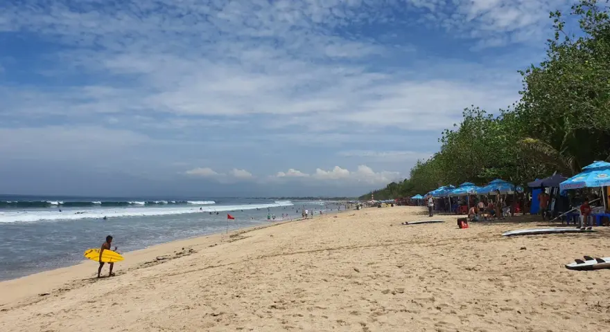 En man med en gul surfbräda går längs Kuta Beach. Vågor rullar in mot stranden där folk solar och surfar.
