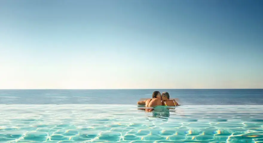 Ett par kysser varandra i en infinitypool med havet i bakgrunden. Perfekt för romantiska resmål.