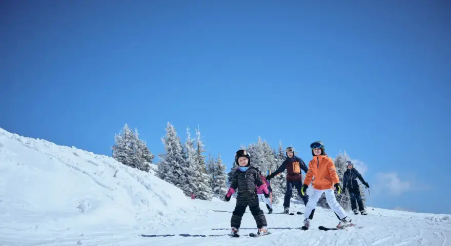 Familj njuter av skidåkning i mars. Barn och vuxna i skidkläder på en snöig backe med träd i bakgrunden. Ett perfekt resmål mars för vinteraktiviteter.