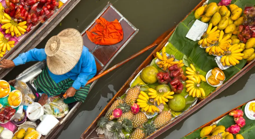 En säljare i en båt på en flytande marknad i Thailand, omgiven av ett överflöd av färsk frukt som bananer, mango och ananas.