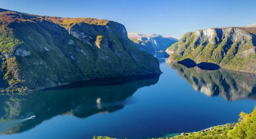 En båt färdas på en spegelblank fjord i Norge, omgiven av gröna bergssluttningar. En tågresa genom detta landskap vore en dröm.