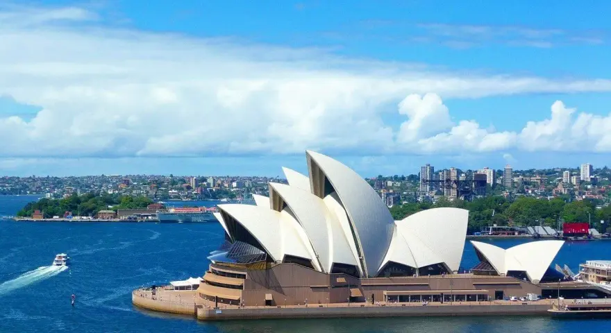 Sydney stad med operahuset i förgrunden och en båt på det blå vattnet.