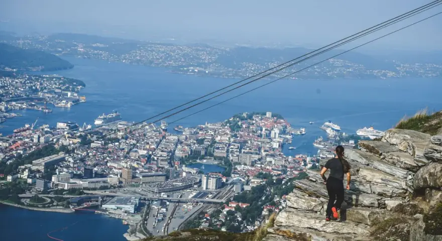 En kvinna står på en klippa och blickar ut över en stad vid havet, med berg i bakgrunden.