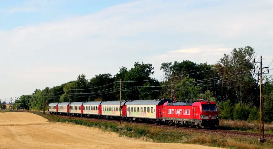 Ett rött lok drar ett långt tåg med vita vagnar genom ett landskap med guldigt höstfält och gröna träd. Kanske är det ett nattåg till Berlin.