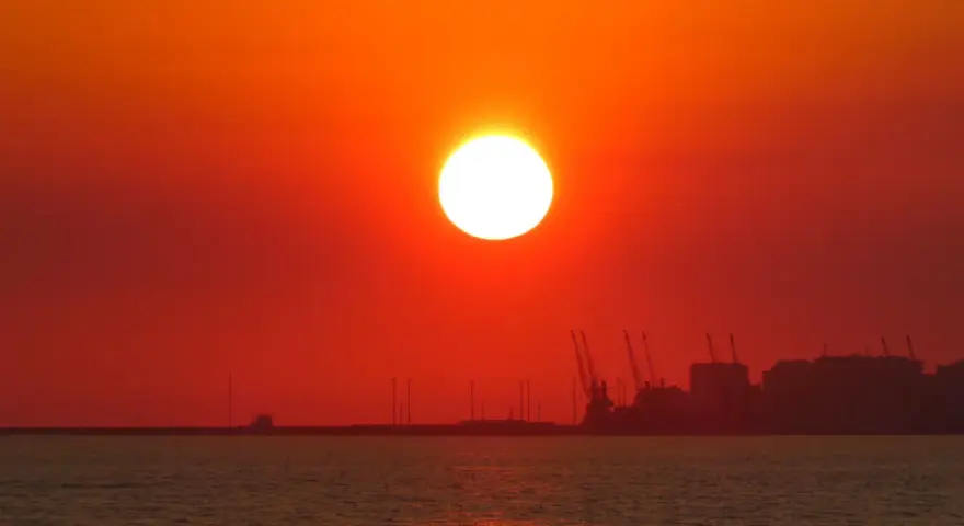 En glödande sol går ner över havet, med silhuetter av hamnkranar och byggnader i fjärran. En av de fina solnedgångar som får en att drömma sig bort.