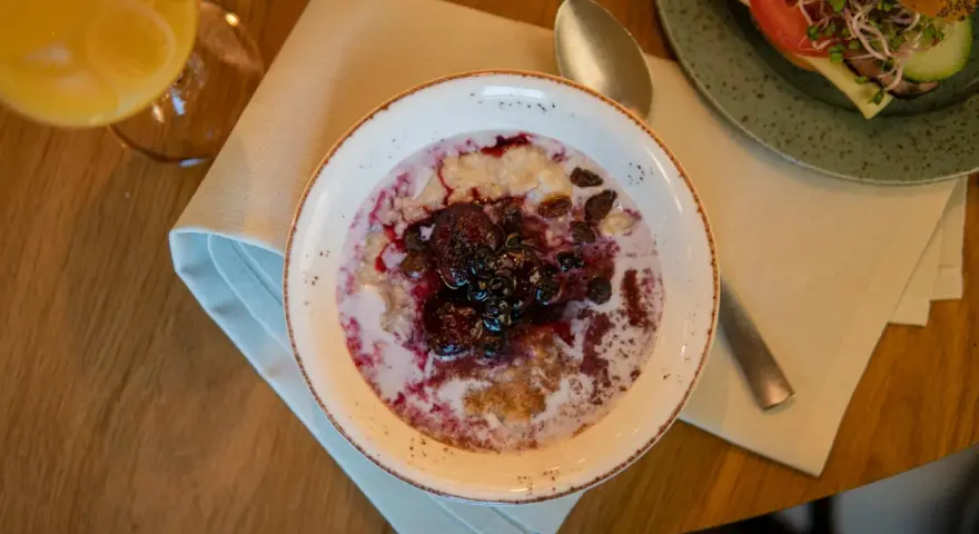 En skål med gröt toppad med bär och russin, en del av en kroatisk frukost. Bredvid syns en smörgås och ett glas juice.