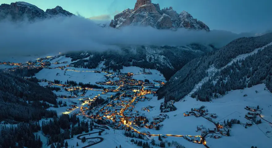 En snötäckt by i Italien på vintern, upplyst av tusentals ljus. Majestätiska berg reser sig bakom, inbäddade i dimma.
