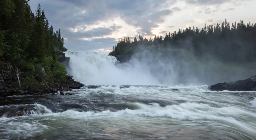 Vackra vattenfall dånar nerför klipporna, omgivna av en tät grön skog under en dramatisk molnhimmel.