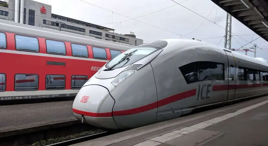 Ett modernt ICE-tåg står redo vid perrongen, perfekt för en resa med tåg till Tyskland. Bredvid syns ett rött dubbeldäckartåg.