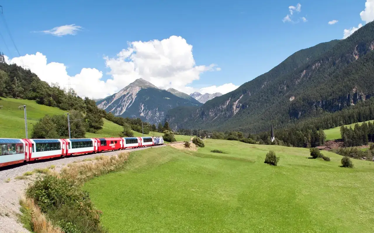 Tåg i alplandskap, perfekt för dina interrail tips. Grönskande ängar, berg och en kyrka.