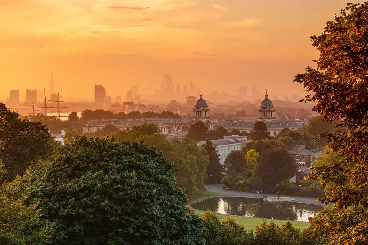 Gyllene solnedg&aring;ng &ouml;ver London. I f&ouml;rgrunden gr&ouml;nska och en damm. I bakgrunden skymtar silhuetten av staden med The Shard och ett segelfartyg. Vissa omr&aring;den att undvika i London kan vara sv&aring;ra att se i denna dimma.