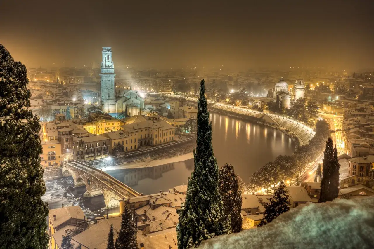 Vacker vy &ouml;ver en sn&ouml;t&auml;ckt stad i Italien p&aring; vintern. En flod slingrar sig genom staden, kantad av upplysta byggnader och tr&auml;d.