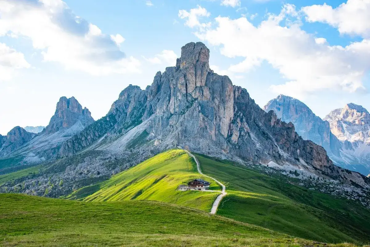 En stig leder till ett hus vid foten av ett majestätiskt italien berg. Grönskande kullar möter dramatiska klippformationer under en blå himmel med moln.