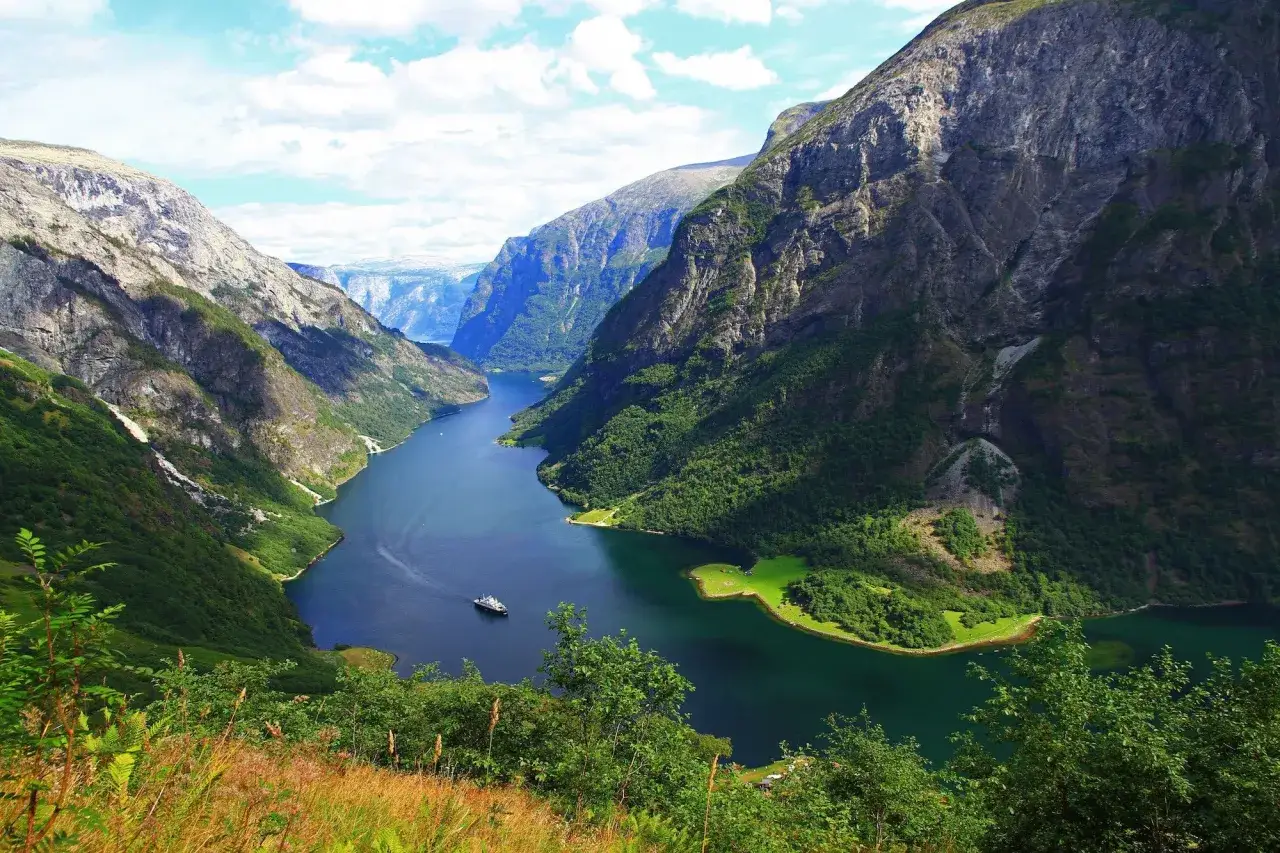 En b&aring;t kryssar p&aring; en djupbl&aring; fjord omgiven av gr&ouml;na berg. En perfekt vy f&ouml;r en semester i Norge.