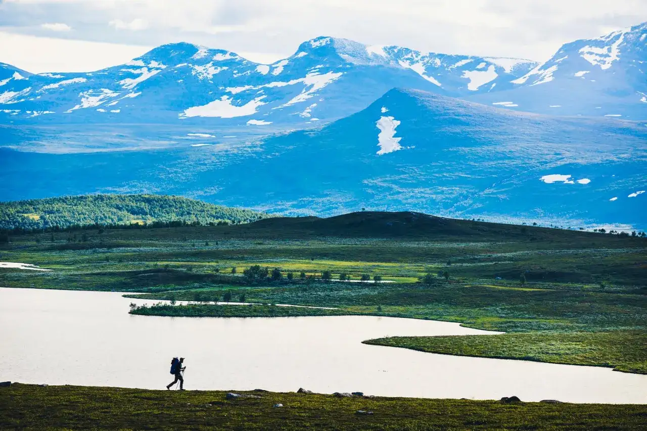 Vandrare med ryggs&auml;ck vid en sj&ouml;, med sn&ouml;kl&auml;dda berg i bakgrunden. Vackert landskap med berg i Sverige.