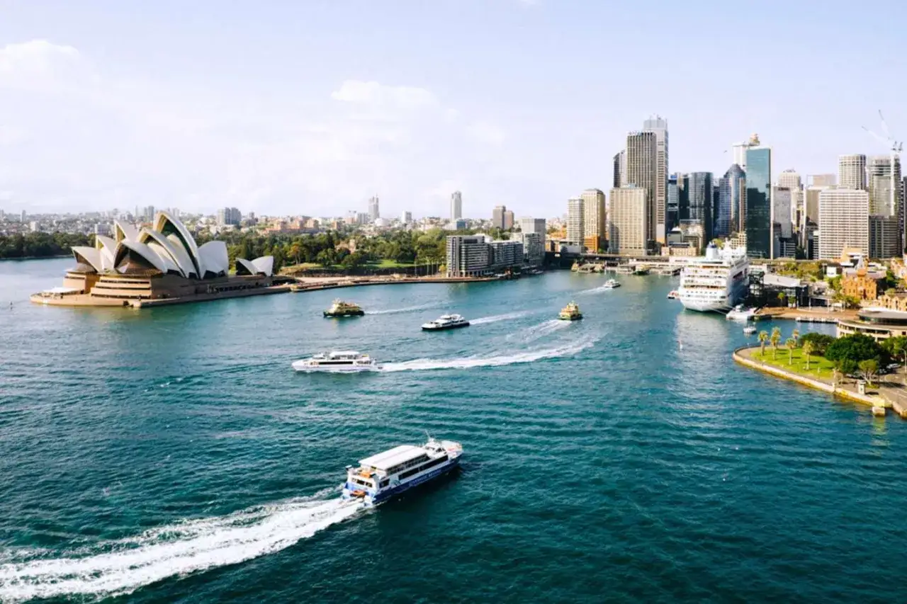 F&auml;rgglad vy &ouml;ver Sydney stad med operahuset, skyskrapor och flera fartyg som kryssar p&aring; det bl&aring; vattnet.