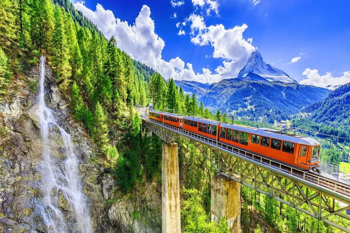 Ett orange t&aring;g korsar en bro &ouml;ver en forsande vattenfall i Schweiz, med majest&auml;tiska berg och en bl&aring; himmel i bakgrunden.
