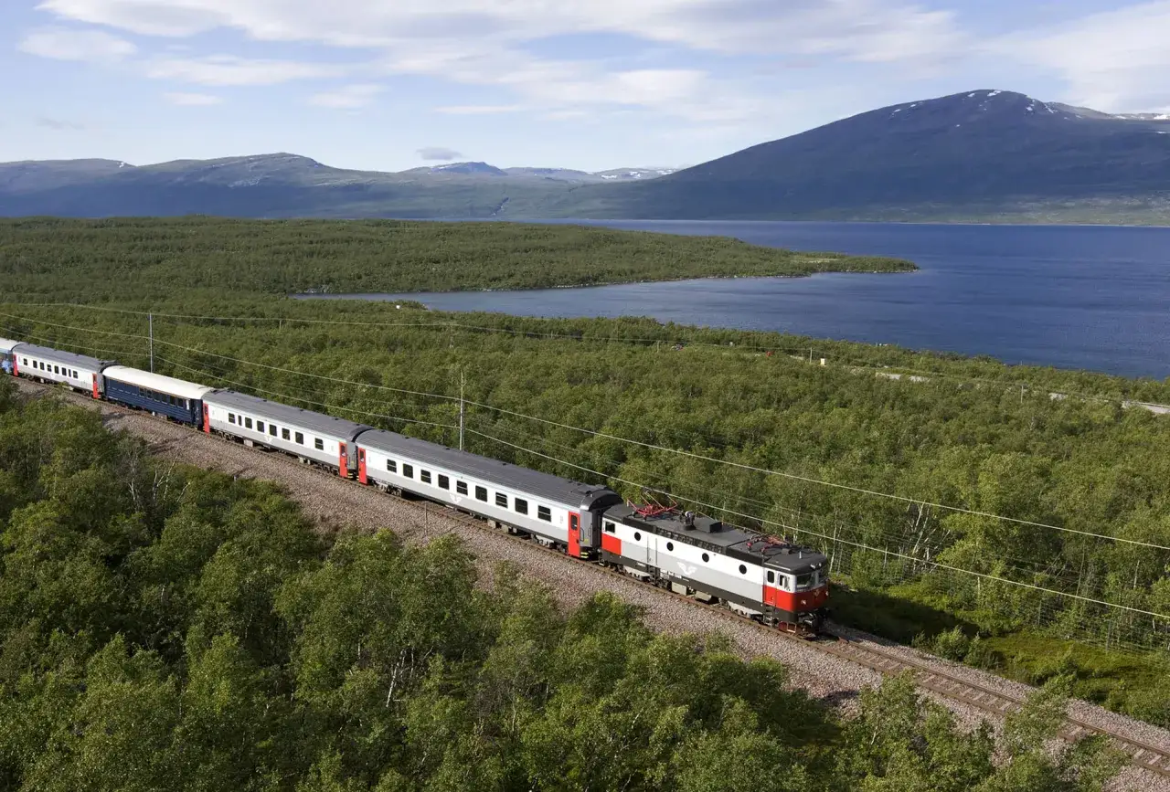 T&aring;g som t&aring;gluffar i Sverige genom gr&ouml;nskande skogar, l&auml;ngs en bl&aring; sj&ouml; med berg i bakgrunden.