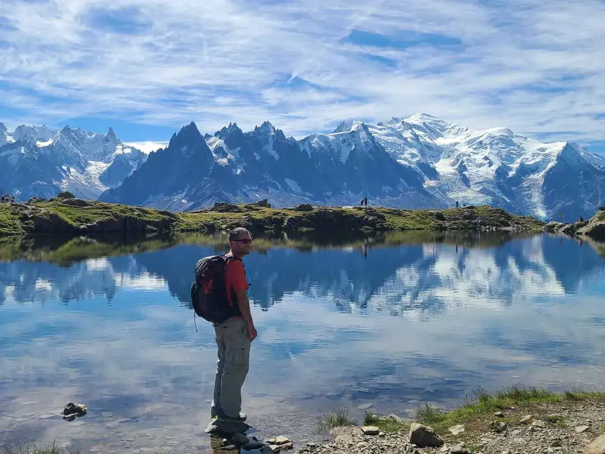 Man med ryggs&auml;ck njuter av en vacker chamonix vandring vid en spegelblank sj&ouml; med sn&ouml;kl&auml;dda berg i bakgrunden.