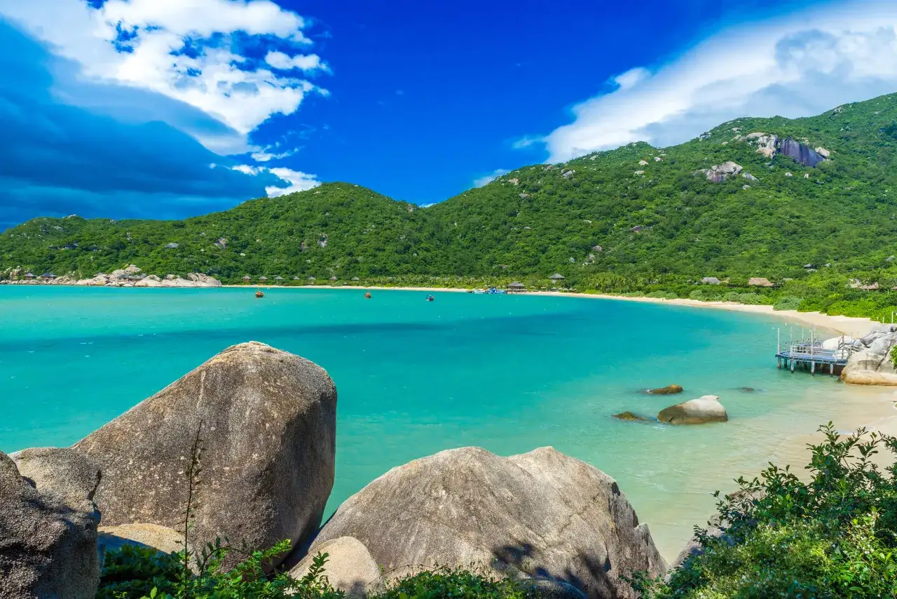 En turkos vik med gr&ouml;na berg och stenar, en strand med sm&aring; hus och en brygga. En idyllisk vy fr&aring;n Nha Trang, Vietnam.