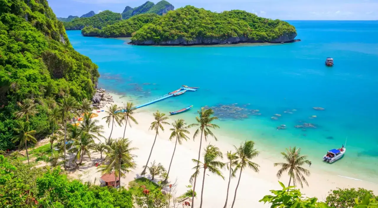 En vit sandstrand med vajande palmer och turkost vatten, en bl&aring; pir med b&aring;tar och gr&ouml;nskande &ouml;ar i bakgrunden. En dr&ouml;msemester p&aring; Koh Samui, Thailand.