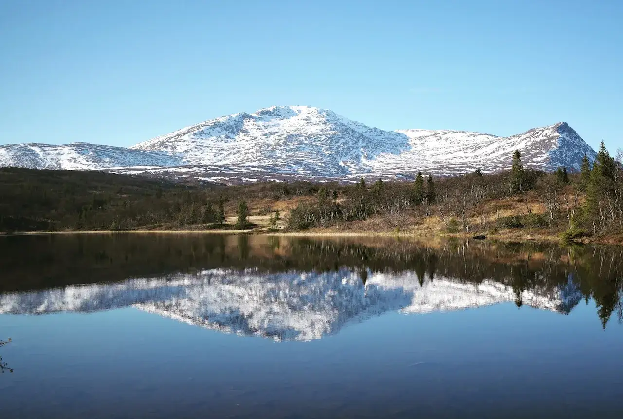 Spegelblank sjö reflekterar snöklädda berg och höstfärgad skog. En perfekt vy för hemester i Sverige.