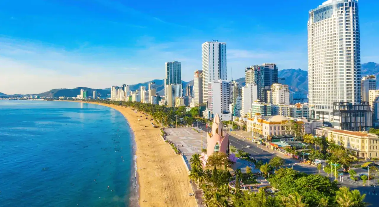 Solig dag vid Nha Trang beach med turkost vatten, gyllene sand och en imponerande stadssilhuett med h&ouml;ga byggnader.
