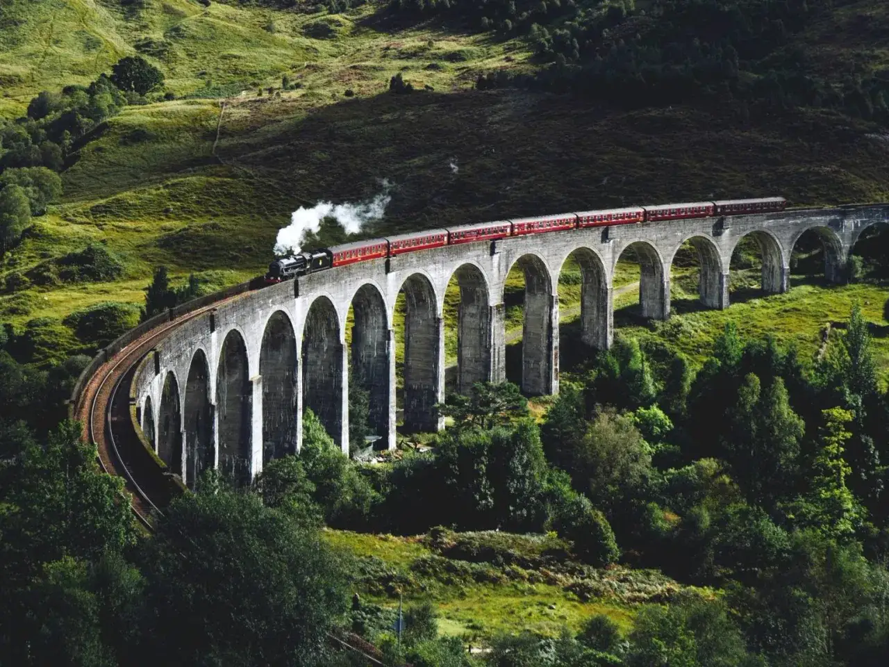 T&aring;g som p&aring;minner om Harry Potter t&aring;g skottland &aring;ngar fram &ouml;ver en storslagen stenbro i ett gr&ouml;nt, kuperat landskap.