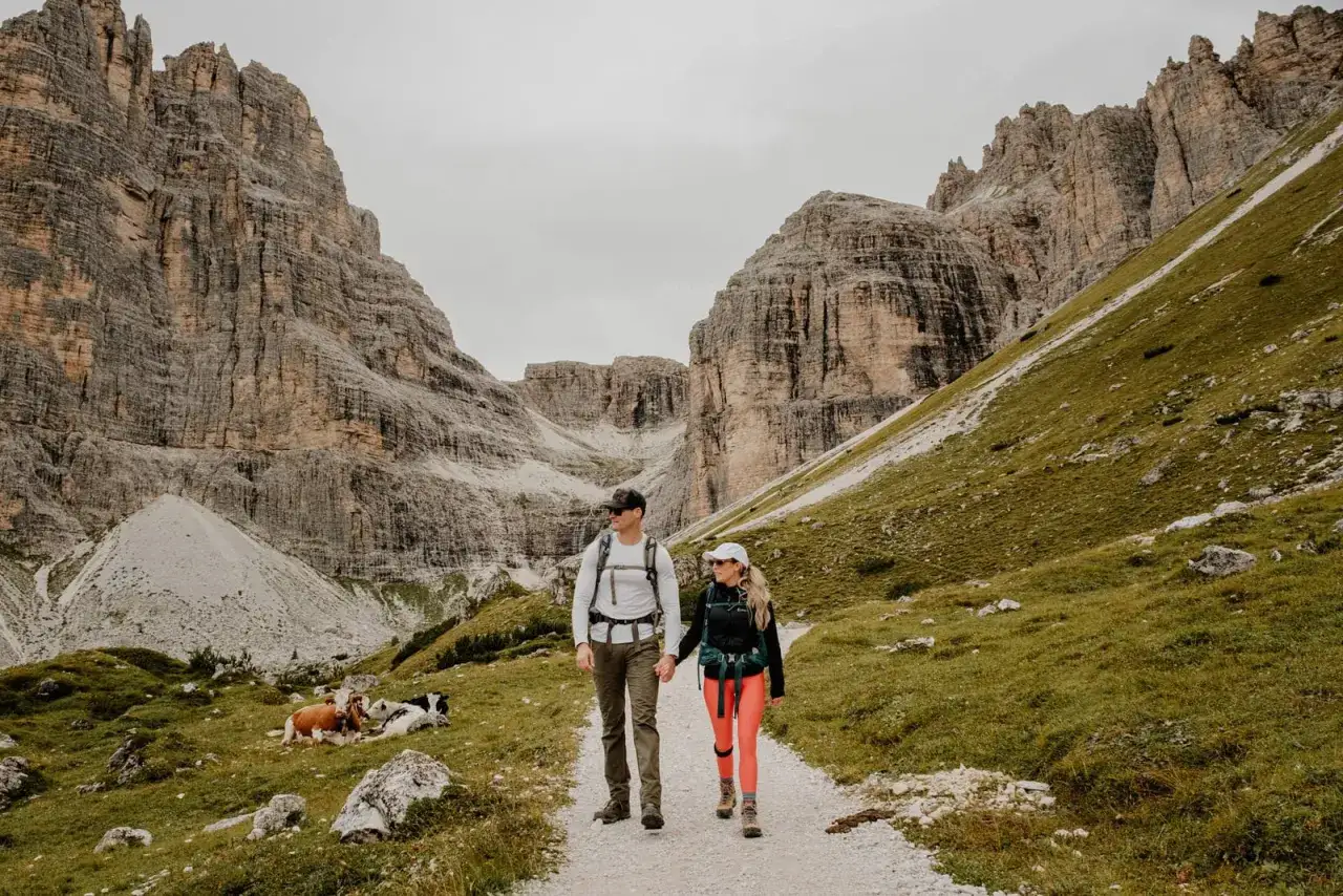 Par som h&aring;ller hand p&aring; en grusv&auml;g under en dolomiterna vandring. Majest&auml;tiska berg reser sig bakom dem, med gr&ouml;na sluttningar och betande kor.