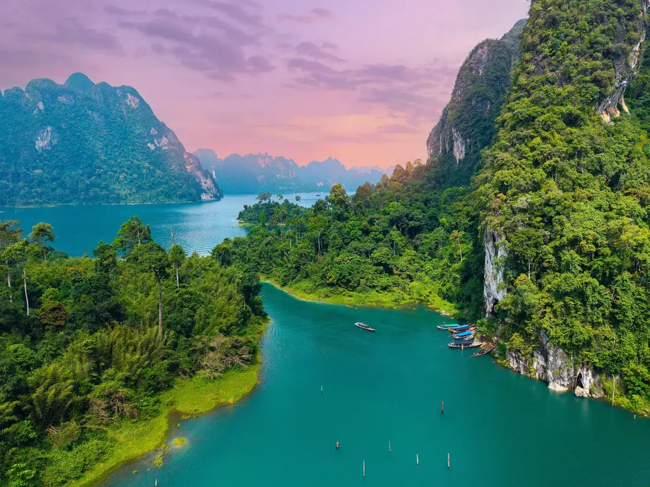 Khao Sok nationalpark med turkost vatten, frodig gr&ouml;nska och dramatiska kalkstensklippor. Sm&aring; b&aring;tar guppar vid en klippv&auml;gg.