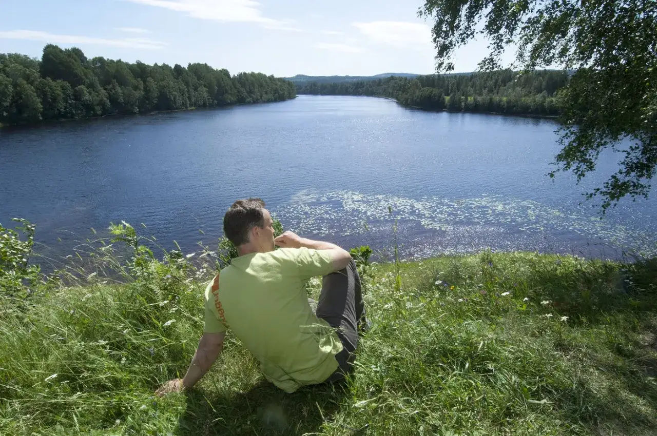 Man sitter p&aring; en gr&auml;skl&auml;dd kulle och blickar ut &ouml;ver en stilla sj&ouml; omgiven av skog. Ett perfekt utflyktsm&aring;l i Dalarna f&ouml;r avkoppling.