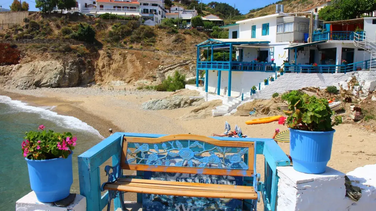 En bl&aring; b&auml;nk med blommor framf&ouml;r en strand i Ikaria, Grekland. En person solar p&aring; stranden.