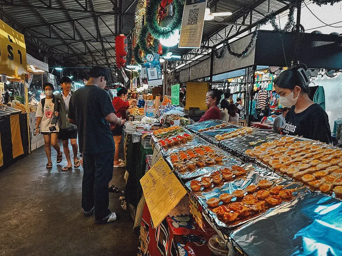 Folk tr&auml;ngs vid matst&aring;nd p&aring; en livlig marknad i Bangkok. Grillspett och sm&aring; stekta &auml;gg ligger uppradade, redo att avnjutas.