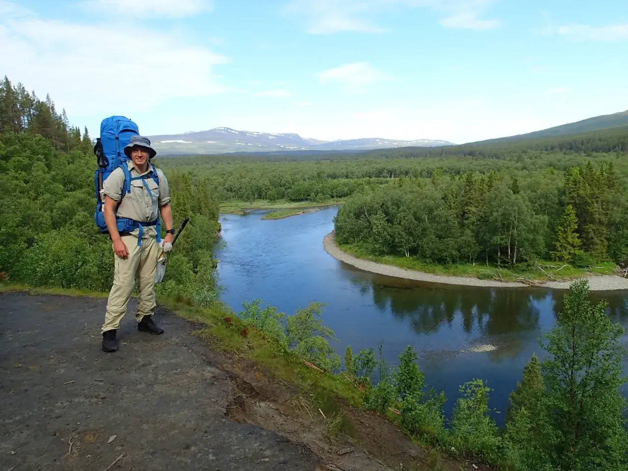 Vandrare med stor ryggs&auml;ck njuter av utsikten &ouml;ver en slingrande flod och fj&auml;llandskap under en v&aring;l&aring;dalen vandring.