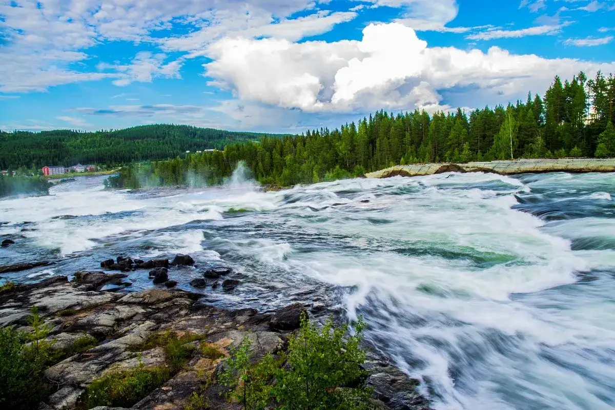 Kraftfullt vattenfall vid Lule&aring;, med forsande vatten, gr&ouml;nskande skog och en bl&aring; himmel med moln.