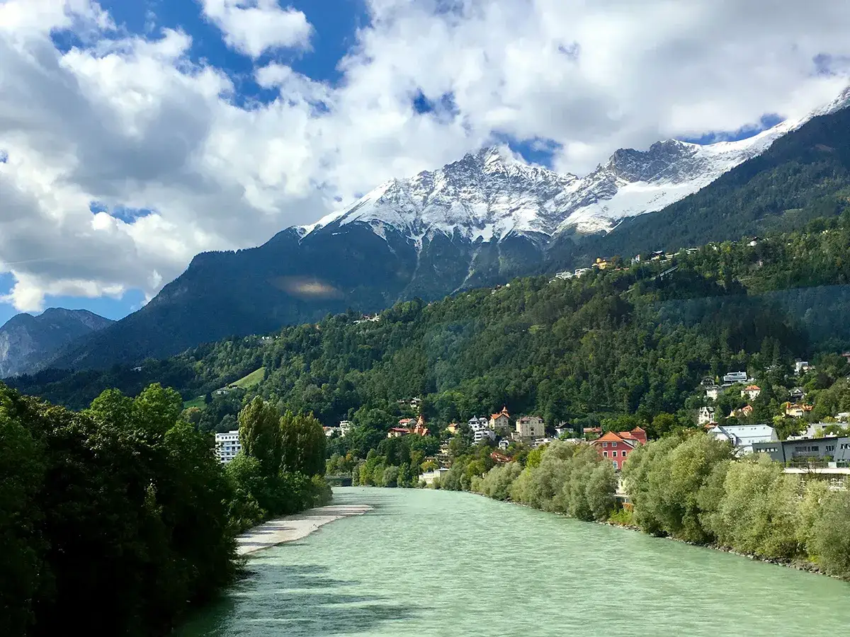 Gr&ouml;n flod flyter f&ouml;rbi ett gr&ouml;nt berg med sn&ouml;kl&auml;dda toppar. Hus kl&auml;ttrar uppf&ouml;r sluttningen, en idyllisk vy som p&aring;minner om Innsbruck skid&aring;kning.
