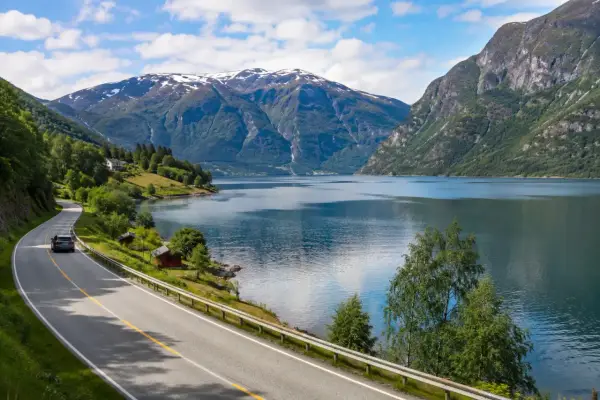 En bil kör längs en slingrande väg vid en fjord, med snöklädda berg i bakgrunden. En perfekt bild för en semester i Norge.
