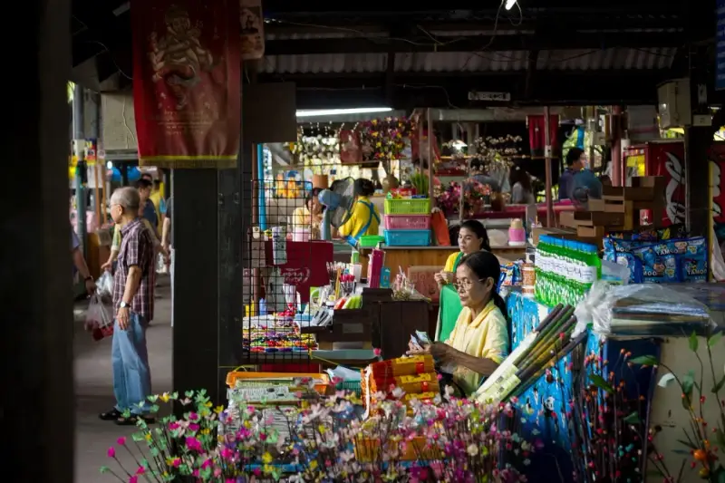 En livlig marknad i Bangkok med försäljare och kunder bland färgglada varor och blommor.