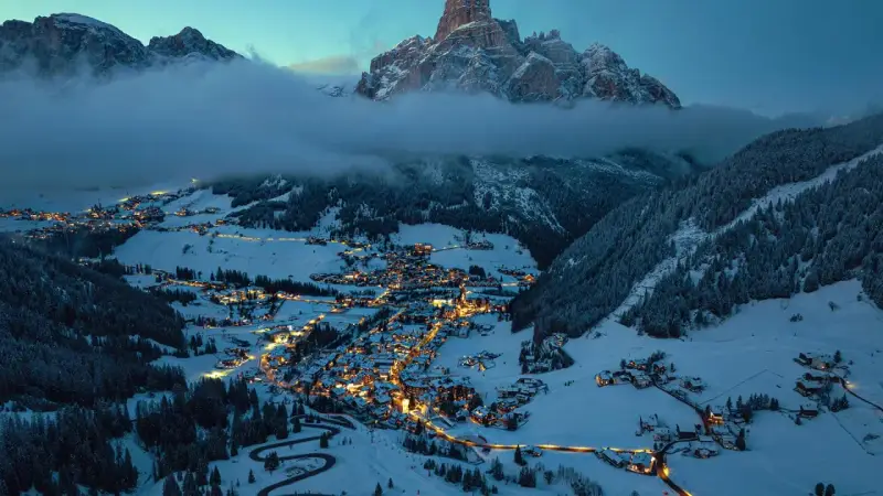 En snötäckt by i Italien på vintern, upplyst av tusentals ljus. Majestätiska berg reser sig bakom, inbäddade i dimma.